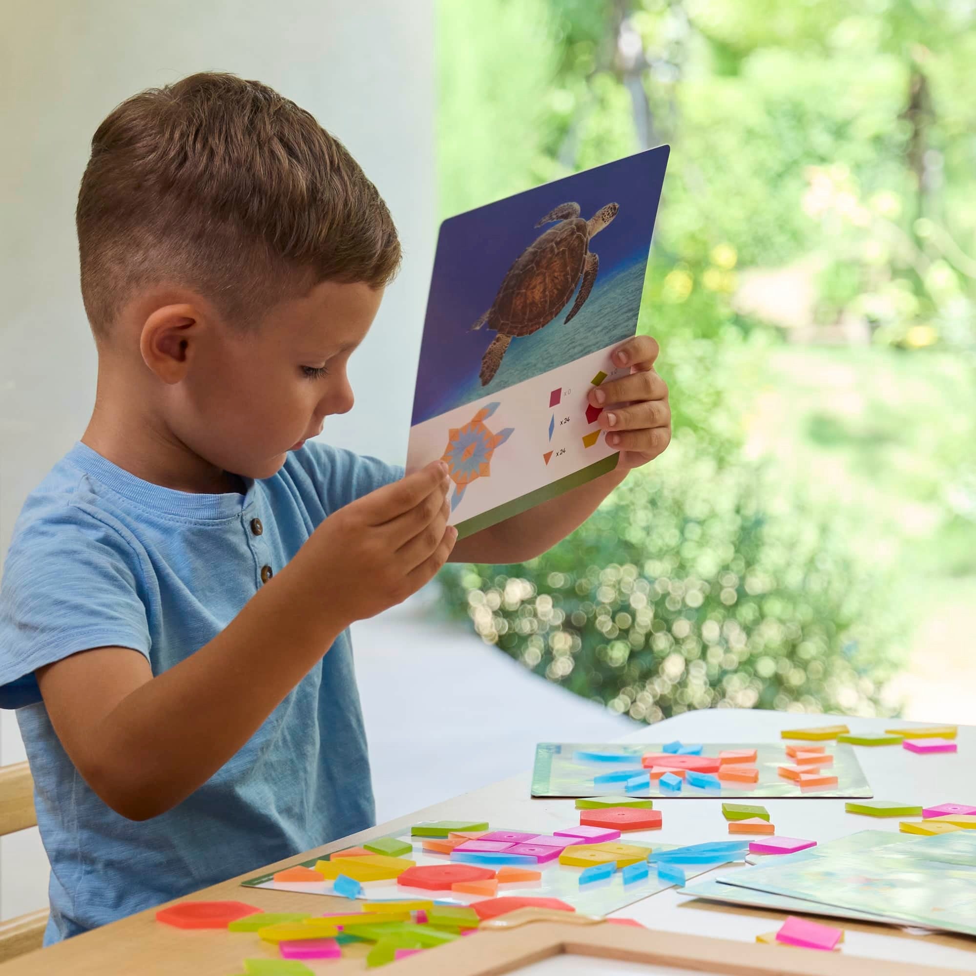 Kind betrachtet Guidecraft durchscheinende Tangram-Fliesen, nutzt Vorlagenkarten zum Legen geometrischer Formen, fördert spielerisch Konzentration, Motorik und kreatives Verständnis im Alltag.kids playing