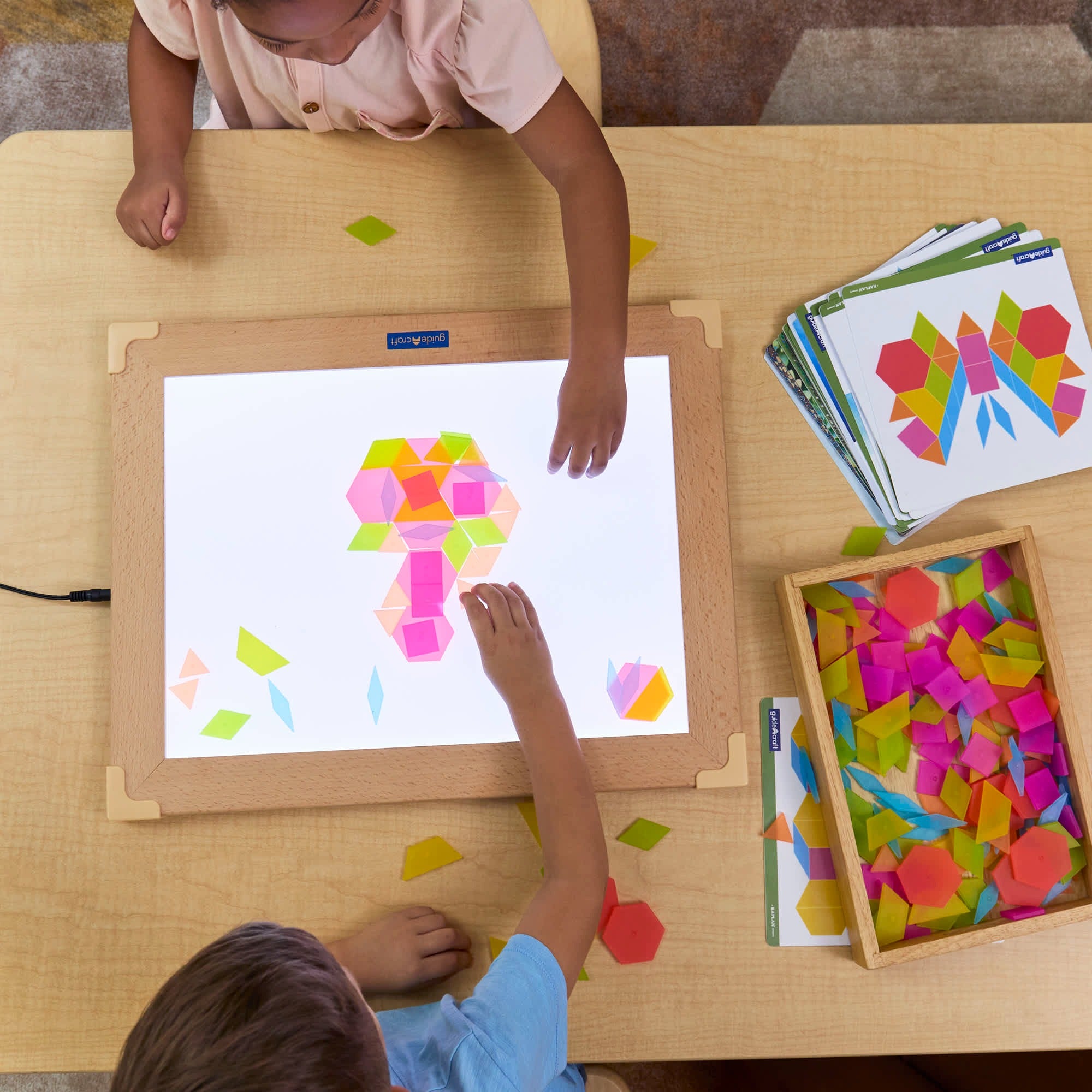 Kinder legen Guidecraft durchscheinende Tangram-Fliesen auf einen Leuchttisch, kombinieren geometrische Formen nach Vorlagen, fördern spielerisch Kreativität, Feinmotorik und mathematisches Verständnis.