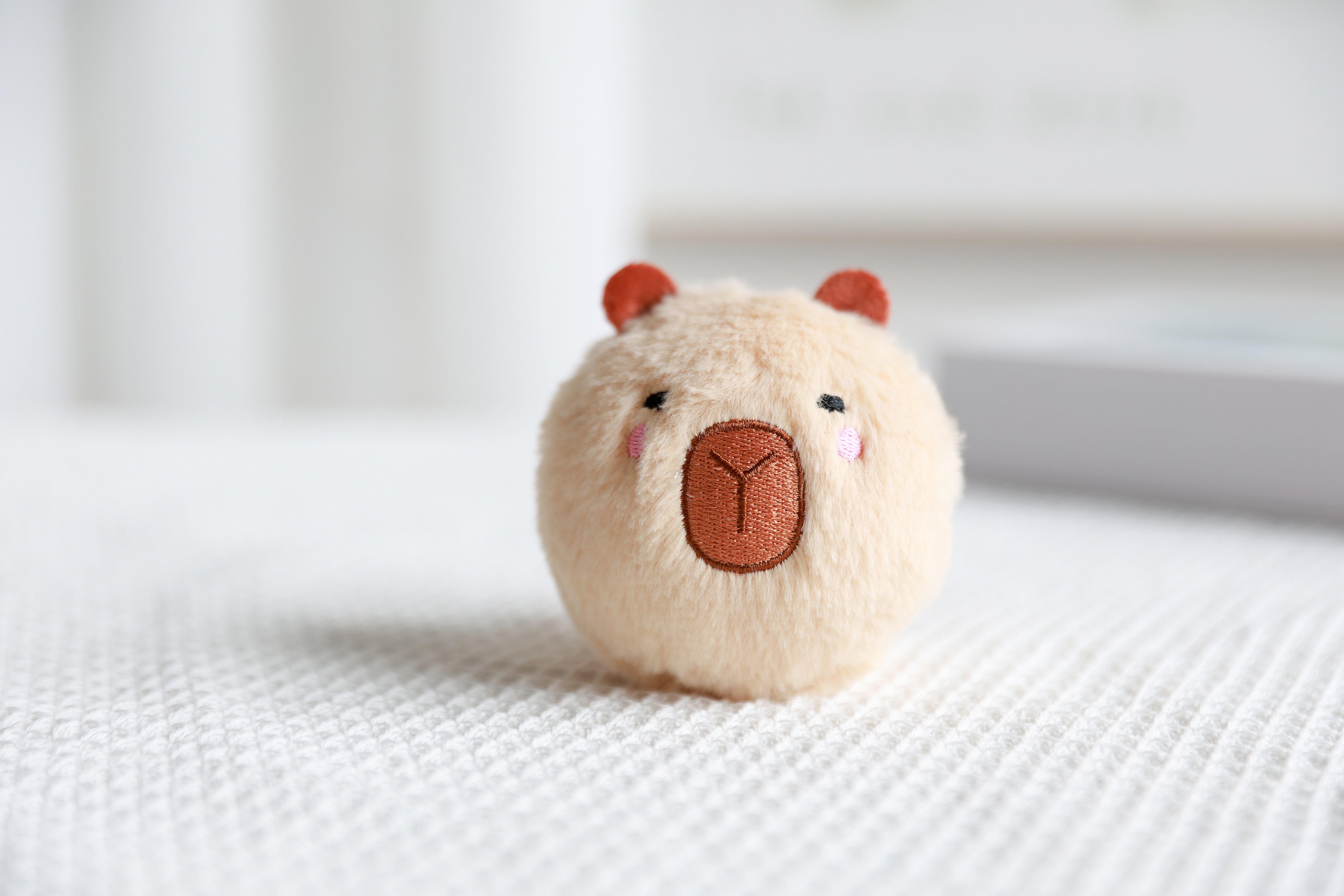 Beiger iTotal Capybara-Plüschball mit weichem Kunstfell als dekoratives Spielzeug auf einer strukturierten weißen Stoffoberfläche.