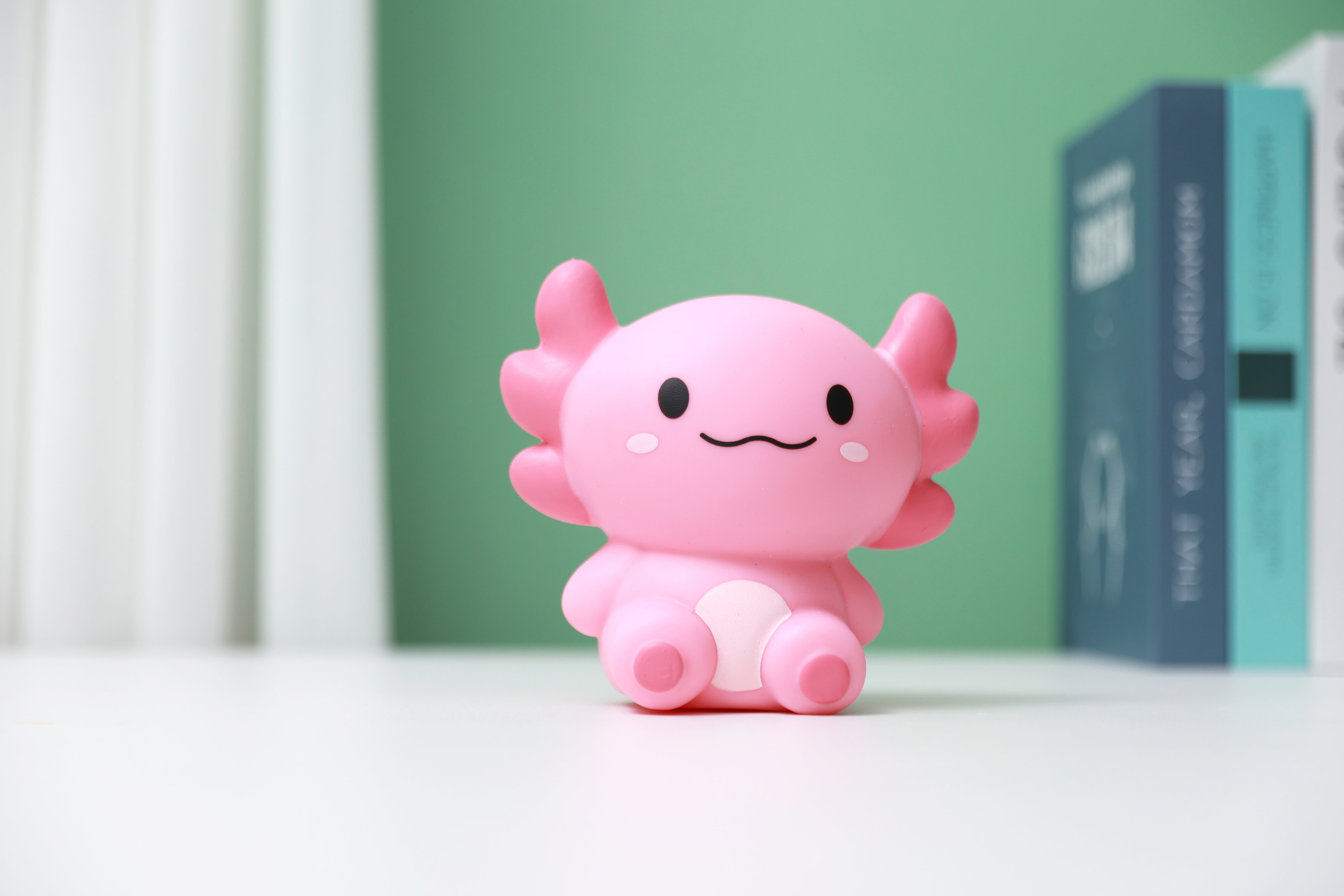 Rosa iTotal Squeeze Axolotl XL als dekoratives Antistress-Spielzeug auf einem Schreibtisch vor grünem Hintergrund.